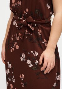 Καφέ φόρεμα με ροζ floral μοτίβο, δεμένο στη μέση με φιόγκο, που αφήνει μέρος του μπράτσου και του χεριού να φαίνεται καθώς ακουμπούν στο πλάι.