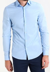 Ljusblå långärmad skjorta med knappdown-krage, slät textur och rundad nederkant. Har vita knappar fram.