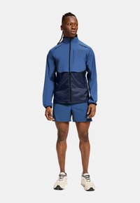 Blue athletic jacket with a navy lower section; smooth texture. Paired with matching blue shorts and white running shoes.
