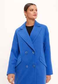 Niebieski dwurzędowy płaszcz z teksturowanej tkaniny z klapami, dwoma przednimi kieszeniami i dopasowanymi guzikami. Gładkie wykończenie i dopasowany krój.