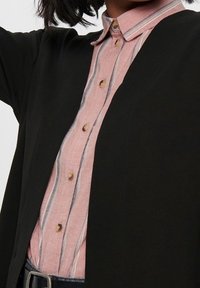 Czarny kardigan noszony na różowo-białą koszulę z guzikami w kolorze złotym. Posiada długie rękawy i gładką fakturę.