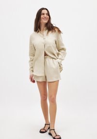 Light beige linen button-up shirt with long sleeves, paired with matching shorts featuring an elastic waistband. Flat sandals complete the outfit.