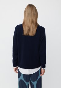 Granatowy sweter z dzianiny, długi rękaw i ściągacz na dole, noszony na białej koszuli, w zestawie z wzorzystymi niebieskimi spodniami z dużymi okręgami.