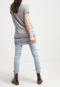 Túnica gris con mangas enrolladas, que presenta una franja horizontal morada en el dobladillo, combinada con jeans azul claro y botas de cordones marrón oscuro.