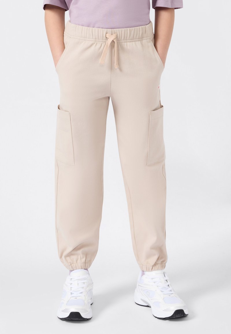 Personne portant un pantalon cargo beige avec des poignets élastiques et un cordon de serrage à la taille, associé à des chaussures de sport blanches et à une chemise à manches courtes lavande.