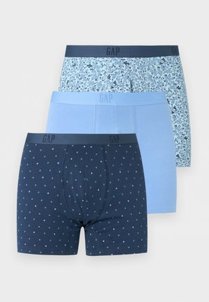 Γυμνασμένος άνδρας με σκούρο δέρμα στέκεται ξυπόλητος φορώντας σκούρα μπλε μποξεράκια με μικρό λευκό πουά σε απλό φωτεινό φόντο.