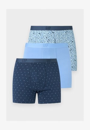 Γυμνασμένος άνδρας με σκούρο δέρμα στέκεται ξυπόλητος φορώντας σκούρα μπλε μποξεράκια με μικρό λευκό πουά σε απλό φωτεινό φόντο.