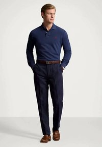 Άνδρας που φοράει ναυτικό μπλε πουκάμισο με μακριά μανίκια, ναυτικό ριγέ παντελόνι με καφέ ζώνη και καφέ παντόφλες, στέκεται με τα χέρια στις τσέπες.