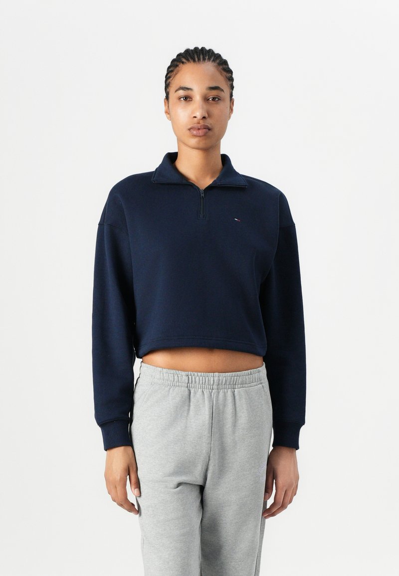 Marine blå cropped sweatshirt med kvart lynlås, ribbet krave og lange ærmer, kombinert med lysegrå joggebukser. Har en liten logo.