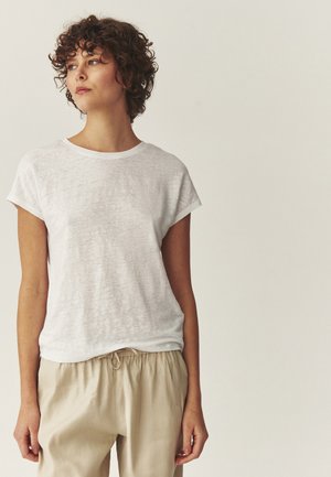 Göndör hajú személy, egyszerű fehér rövid ujjú inget és bézs színű, húzózsinóros nadrágot visel, oldalra néz neutrális háttér előtt.