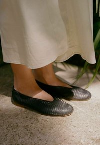 Scarpe nere basse con un motivo testurizzato simile al coccodrillo, punta arrotondata e suola flessibile marrone, posizionate su una superficie in cemento.