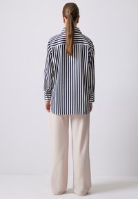 Camicia a righe blu navy e bianca, a maniche lunghe con chiusura a bottone, abbinata a pantaloni beige chiaro a gamba larga, vista da dietro.