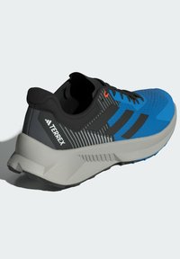 Blå och svart Adidas Terrex trail löparsko med grå sula och svarta snören, vinklad för att visa sid- och bakdetaljer.