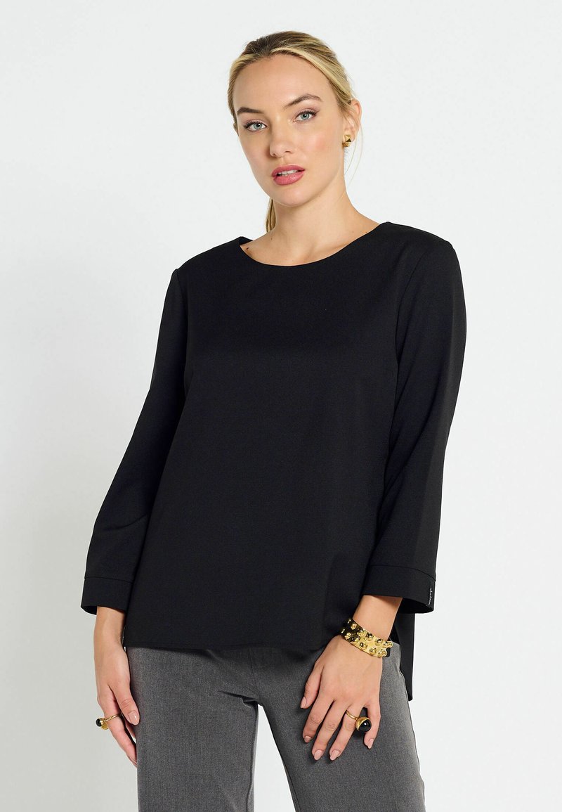 Maglietta nera a maniche lunghe con scollo rotondo, texture liscia e spacchi laterali. Abbinata a pantaloni grigi, con accenti di gioielleria dorata.