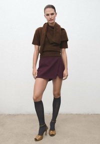 Donna con un maglione corto a maniche corte marrone abbinato a una sciarpa, una mini gonna bordeaux, calze nere fino al ginocchio e sandali aperti color beige.