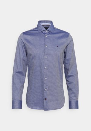 Pánská modrá košile s dlouhým rukávem, knoflíkovým zapínáním vpředu, špičatým límečkem a malým logem na levé straně hrudi na bílém pozadí.