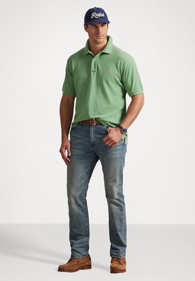 Άνδρας που φοράει πράσινο polo shirt, μπλε τζιν, καφέ ζώνη και παπούτσια, ναυτικό καπέλο με την ένδειξη "Ralph" και ρολόι χειρός, στέκεται με το ένα χέρι στην τσέπη.