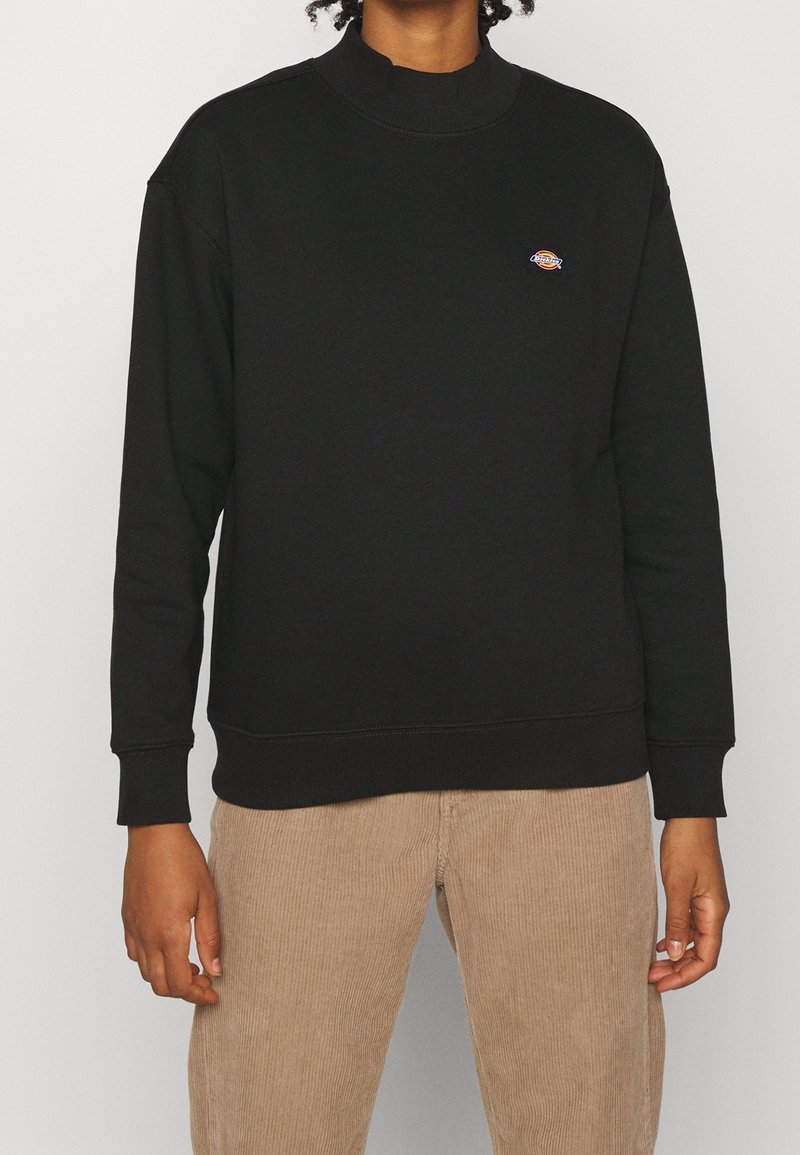Personne portant un pull noir à manches longues avec un petit logo coloré sur la poitrine et un pantalon en velours beige, debout devant un fond uni.