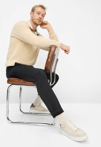 Sweat-shirt beige à col zippé, pantalon noir, baskets crème avec détails texturés, assis sur une chaise chromée avec coussin en cuir marron.