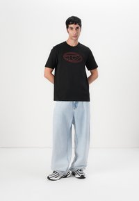 Svart kortermet t-skjorte med rødt ovalt logo på fronten, kombinert med lyseblå oversized jeans og svarte og hvite mønstrede joggesko.