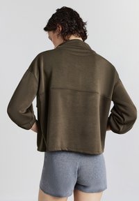 Una persona che indossa una maglietta lunga verde oliva larga e pantaloni corti di maglia grigia, in piedi con le mani sui fianchi, girata di spalle contro uno sfondo neutro.