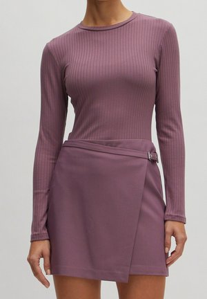 Vrouw die een mauve geribbelde top met lange mouwen en een bijpassende wikkelrok met zijkant gesp draagt, staand tegen een effen achtergrond.