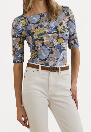 Haut à motif floral en bleu, jaune et brun, avec un col droit et des manches mi-longues jusqu'aux coudes ; associé à un jean blanc et une ceinture marron.