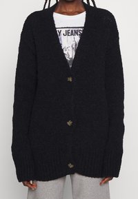 Person, der einen schwarzen, übergroßen, knöpfbaren Cardigan über einem weißen T-Shirt mit Grafik und hellgrauen Hosen trägt, von den Schultern bis zur Hüfte gezeigt.