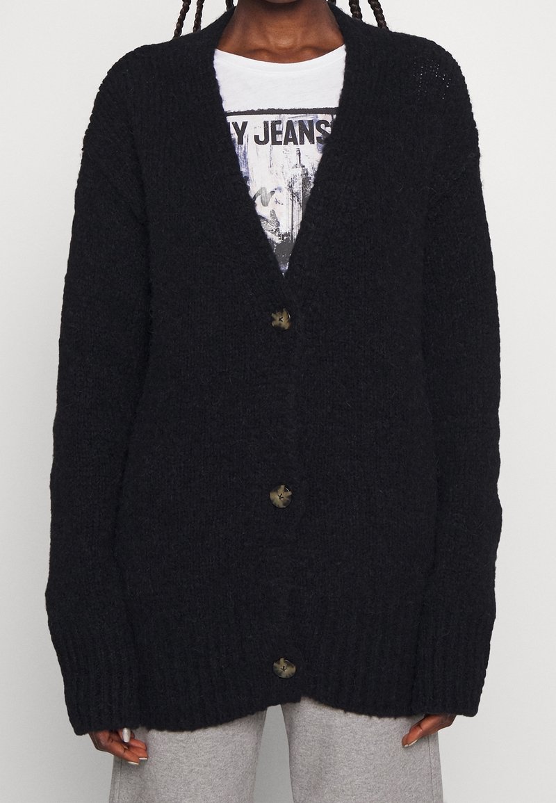 Person, der einen schwarzen, übergroßen, knöpfbaren Cardigan über einem weißen T-Shirt mit Grafik und hellgrauen Hosen trägt, von den Schultern bis zur Hüfte gezeigt.