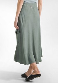 Groene, tot op de grond vallende rok met een gladde textuur, voorzien van een geplooide taille en zijzak. Gecombineerd met zwarte instapschoenen.