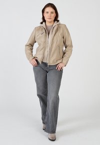 Beige läderjacka med zippered front, kviltade detaljer och avtagbar huva, tillsammans med grå vida jeans och ljusa stövlar.