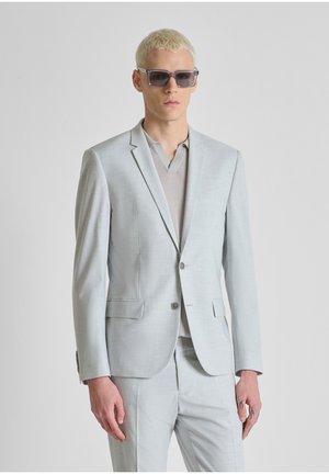 Jonge man met kort blond haar, grijze zonnebril, een lichtgrijs colbert, bijpassende broek en een beige overhemd met kraag.