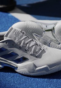 Biele tenisky Adidas s modrými pruhmi a reflexnými striebornými detailmi na modrom textúrovanom povrchu.