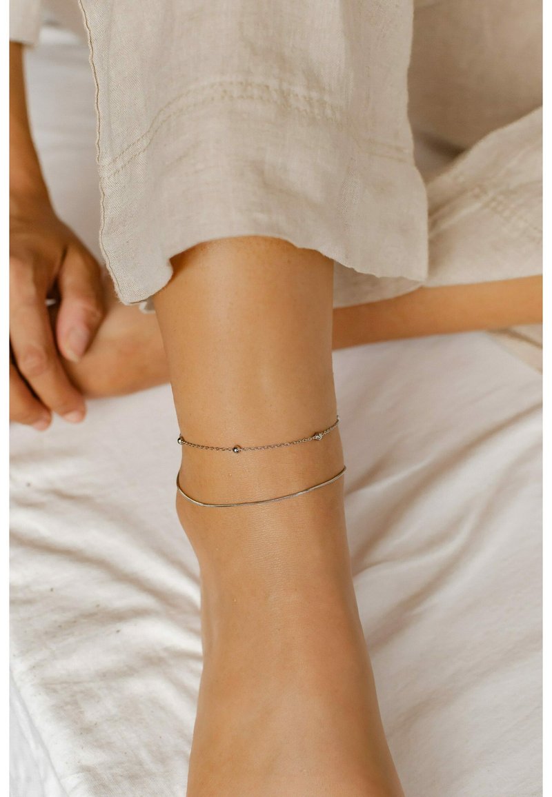 Delgadas pulseras de tobillo de plata, una con pequeñas cuentas, superpuestas en un tobillo bronceado. Un suave fondo de tela en tonos neutros realza el contraste.