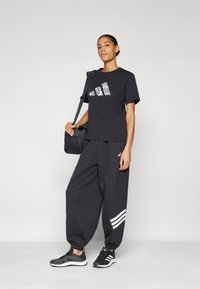 Černý sportovní outfit, který obsahuje tričko s texturovaným bílým logem a volné tepláky se bočním proužkem. Celý vzhled doplňují černé tenisky.