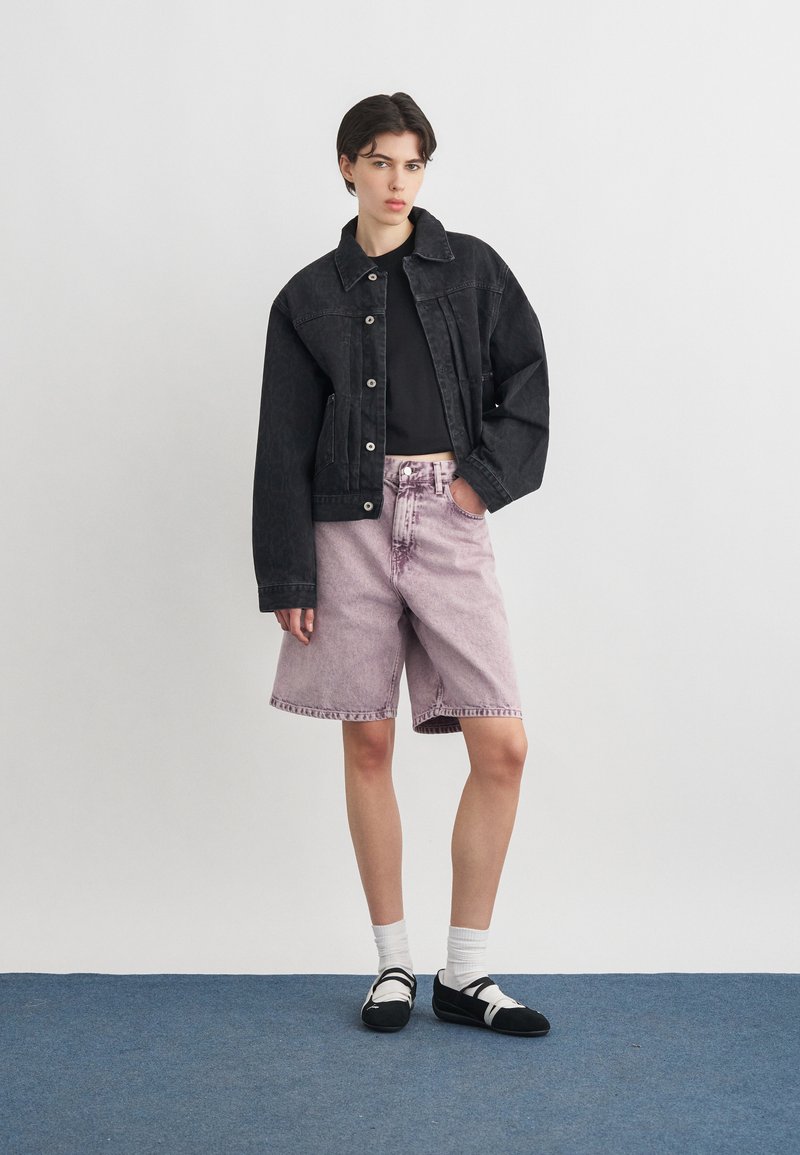 Giovane in piedi che indossa una giacca di jeans nera, top nero, pantaloncini lilla chiaro fino al ginocchio, calzini bianchi e scarpe nere su sfondo semplice.