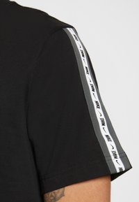 Černé tričko s krátkým rukávem, se šedým páskem podél ramenních švů, zdobené opakujícím se bílým textem "Nike". Hladká textura.