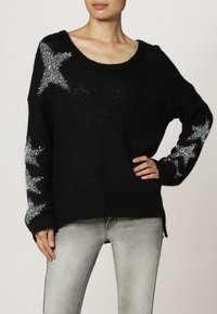 Pull en tricot noir avec un décolleté large et des manches longues, ornée d'accents d'étoiles argentées sur les manches. Texture douce, coupe décontractée.