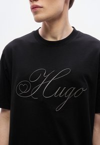 Μαύρο βαμβακερό μπλουζάκι με μεταλλικό ασημί χειρόγραφο λογ ότυπο "Hugo" στο μπροστινό μέρος. Απλός στρογγυλός λαιμός και άνετη εφαρμογή.