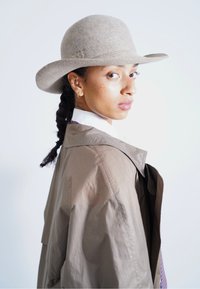 Gray felt hat, loose brown jacket, white collar shirt, and braided hair. Side profile with skin freckles, subtle jewelry accents, and soft lighting.