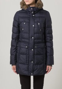 Giacca puffer blu navy con cappuccio in pelliccia sintetica, bottoni a pressione, due tasche anteriori e trapuntatura testurizzata su tutta la superficie.