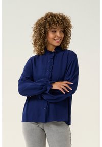 Mörkblå knappad blus med volangkrage och långa puffärmar. Slät tyg med en något lös passform, kombinerad med grå jeans.