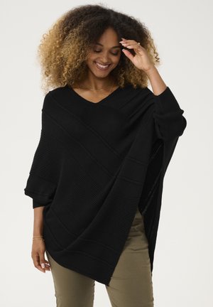 Black oversized knitted poncho with a V-neck, featuring textured ribbed patterns and wide sleeves, worn over olive green pants.