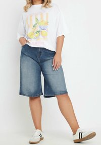 Valkoinen ylisuurikokoinen t-paita, jossa sitruunakuosi, yhdistetty sinisiin farkkubermudoihin. Malli käyttää valkoisia lenkkareita, joissa on mustat raidat.