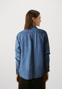 Denim shirt in medium blue, featuring long sleeves, buttoned cuffs, a back pleat detail, and a curved hemline.