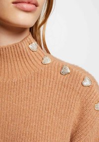 Beige gebreid vest met een hoge ribbelkraag, versierd met glanzende, met kristallen bedekte hartaccenten langs de schouders.