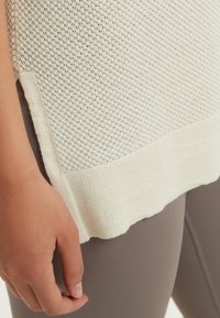 Närbild på en hand som vilar vid fållen på en vit texturerad stickad tröja över taupefärgade leggings.