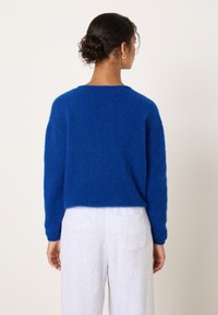 Moteris su tamsiais susuktos kuodos plaukais, dėvinti mėlyną pūkuotą megztinį ir šviesiai pilkas kelnes, stovinti priešais neutralią paprastą foną.