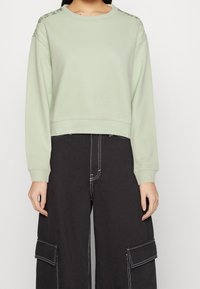 Felpa cropped verde chiaro con maniche lunghe, caratterizzata da dettagli testurizzati sulle spalle. Abbinata a pantaloni cargo neri.
