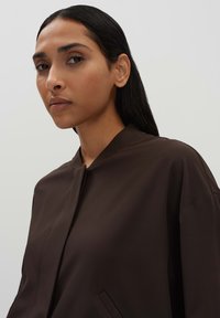 Brown jacket with a structured collar, smooth fabric, and casual design. Features a discreet front fastening and side pockets.